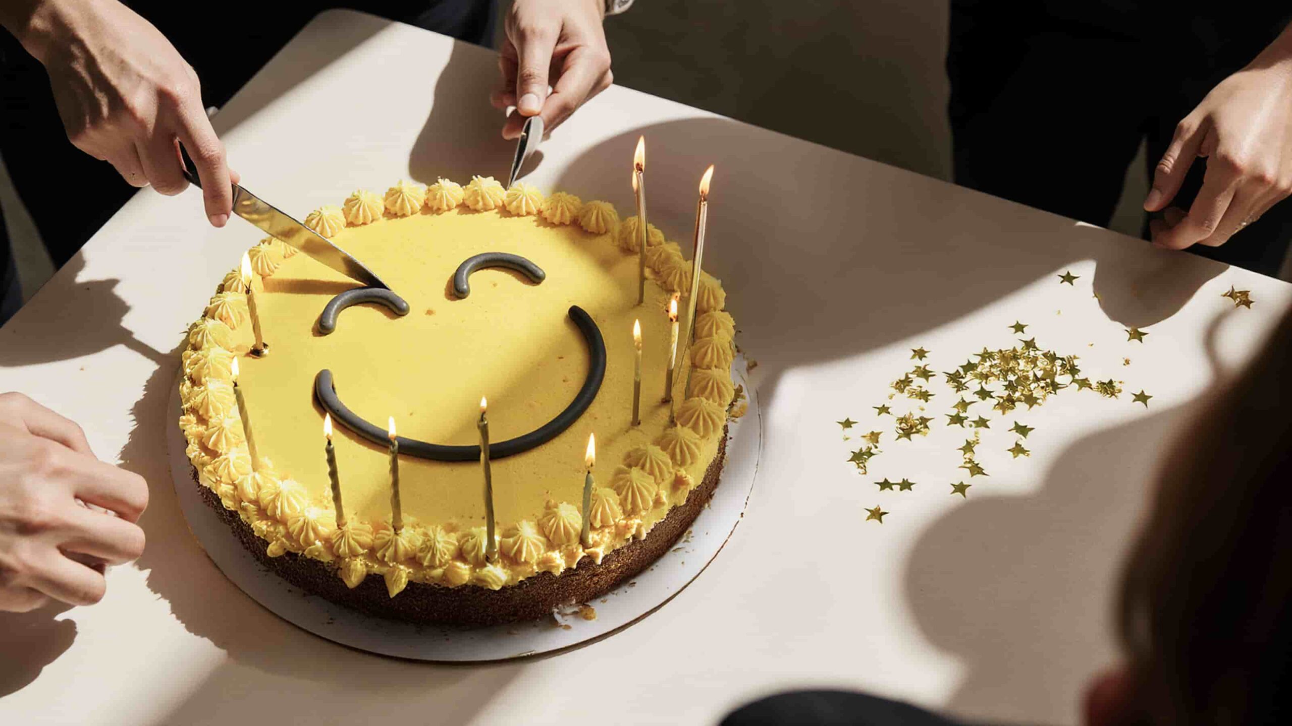 Image of a yellow cake with a smiley face and candles being cut on a white table